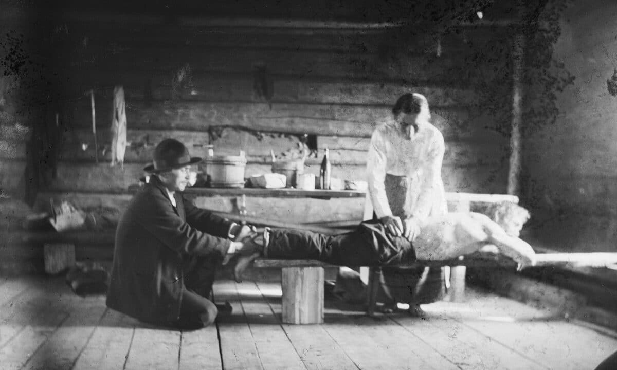 Parantaja apulaisineen hoitaa kolottavaa lonkkaa Taivalkosken Loukonvaarassa. Samuli Paulaharju, 1917. Museovirasto.
